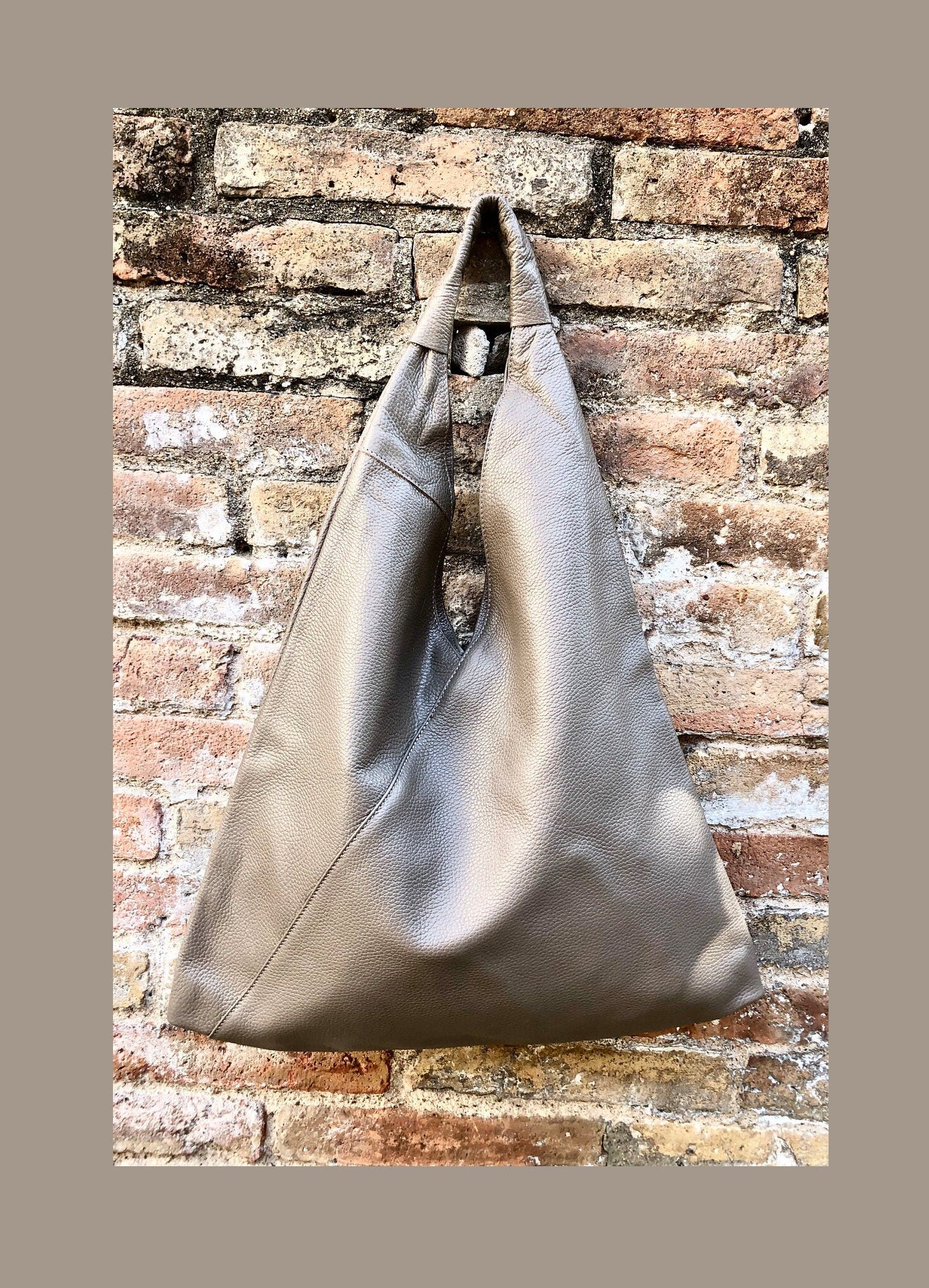 May include: A taupe-colored leather hobo handbag hangs against a brick wall. The bag is triangular in shape with a simple design.  The leather appears soft and supple. This stylish shoulder bag is perfect for everyday use.