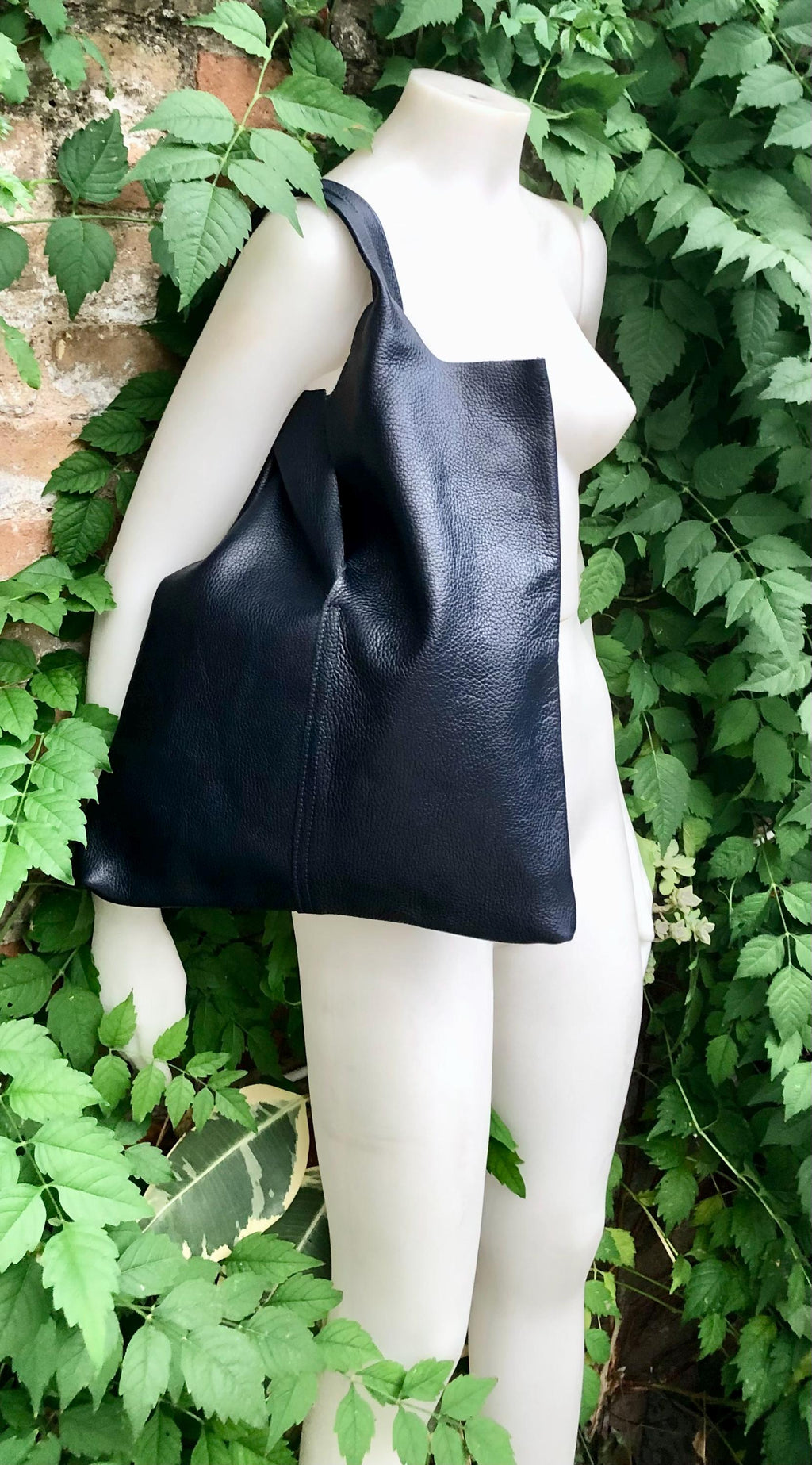 May include: A large, navy blue tote bag with a textured surface. The bag is held by a mannequin and has a simple, rectangular shape with a single shoulder strap. The background features lush green foliage and a brick wall, providing a natural setting for the bag.
