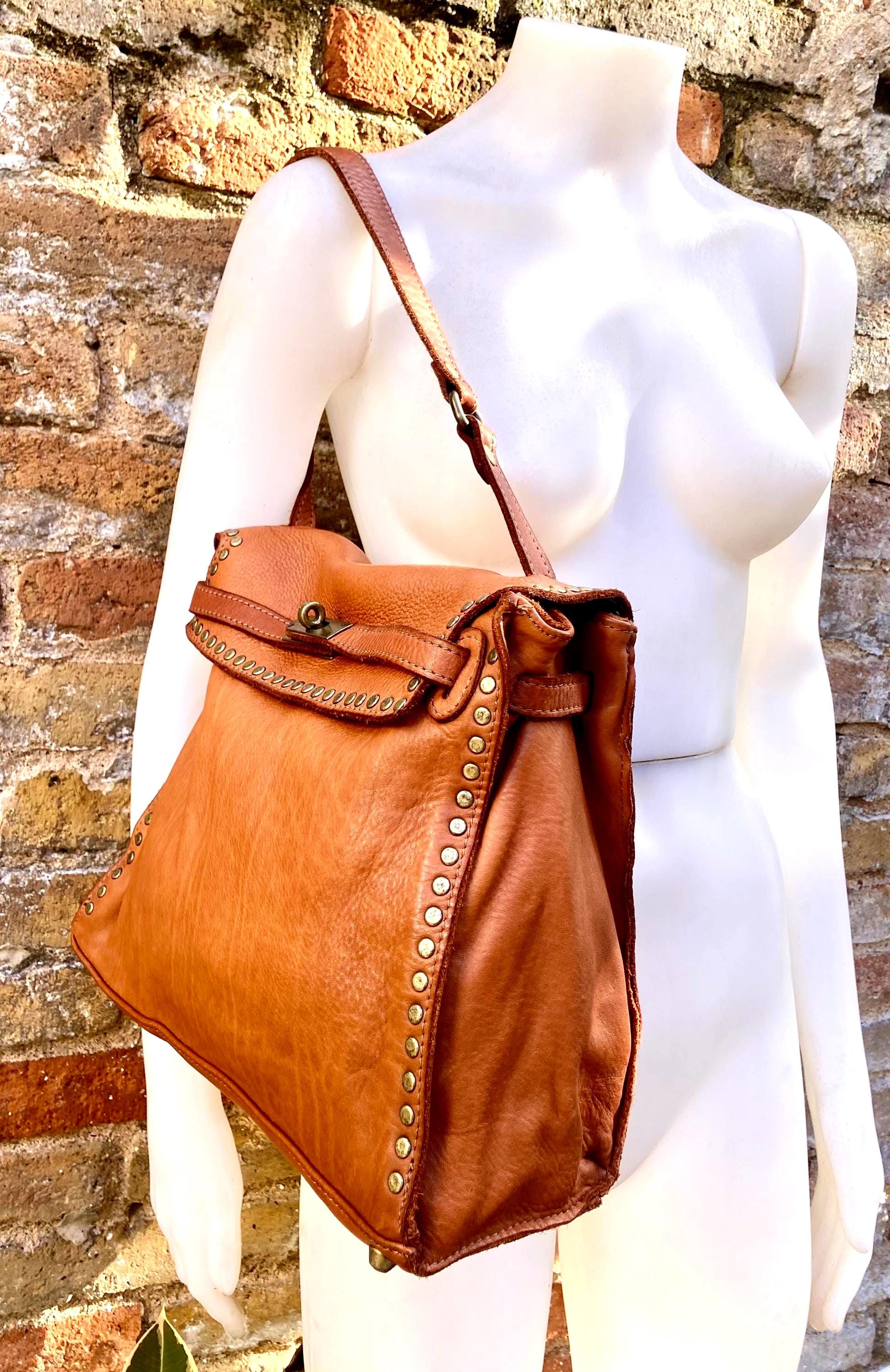 May include: A brown leather shoulder bag with a flap closure and a long strap. The bag is adorned with brass-colored studs along the edges and features a decorative stitched design on the flap. The bag is displayed on a white mannequin.