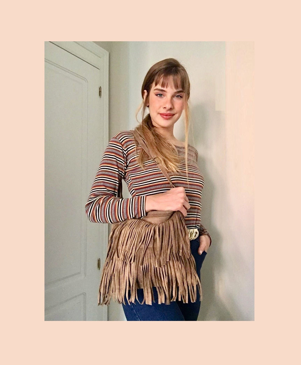 May include: A brown suede fringe bag with a long strap. The bag is worn by a person with long brown hair wearing a striped long-sleeved shirt and blue jeans.
